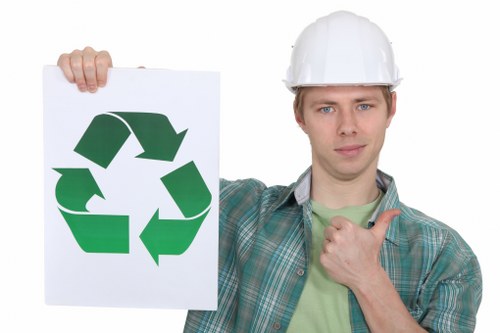 A young man wearing a white safety helmet and a green plaid shirt over a light green T-shirt is standing outdoors, holding a large white sign displaying the green recycling symbol prominently in front of him. His facial expression is neutral, and he is giving a thumbs-up gesture with his right hand, positioned near his chest. Behind him, the background appears out of focus, suggesting a typical outdoor environment. This image visually aligns with waste management and recycling themes relevant to commercial rubbish removal services, such as those provided by Commercial Waste Virginia Water, located locally near Virginia Water in Surrey. The setting emphasizes responsibility for sustainable waste disposal, which complements the company's service offering in the town and surrounding areas.