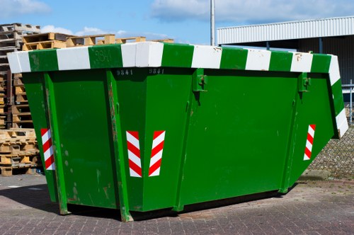 A large, industrial green skip with a robust metal construction and a smooth, painted finish, situated outdoors on a paved driveway. The skip's dimensions are broad and rectangular, with a slight taper at the top opening, which is reinforced by a white and green striped safety border along the upper edge. Red and white reflective hazard decals are affixed to the front and sides, enhancing visibility. In the background, there are stacks of wooden pallets positioned to the left, and a metal building with a corrugated roof to the right, indicating a commercial or industrial environment. Overhead, a tall streetlight or utility pole extends into the sky, which appears partly cloudy, suggesting daytime lighting conditions. The scene suggests a typical site for waste collection and disposal services, with Commercial Waste Virginia Water managing the removal of debris or rubbish from nearby premises, consistent with their local operations in the Virginia Water area, inferred from the contextual clues of the postcode and service relevance to rubbish removal in the region.