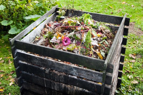 A black wooden composter situated on a grassy area in an outdoor garden setting, filled with a mixture of organic waste including plant trimmings, leaves, and flowers. The compost contains various textures and natural colours such as green, brown, and some pink blossoms. Surrounding the composter are green plants and a lawn, indicating a typical garden environment in Virginia Water. The compost material appears to be in a state of decomposition with visible organic matter and some fresh plant material on top. The scene is well-lit with natural daylight, emphasizing the textures of the compost and the wooden structure, which shows natural weathering. The image reflects the context of waste management and composting, aligning with the services provided by Commercial Waste Virginia Water for rubbish removal and garden waste disposal.