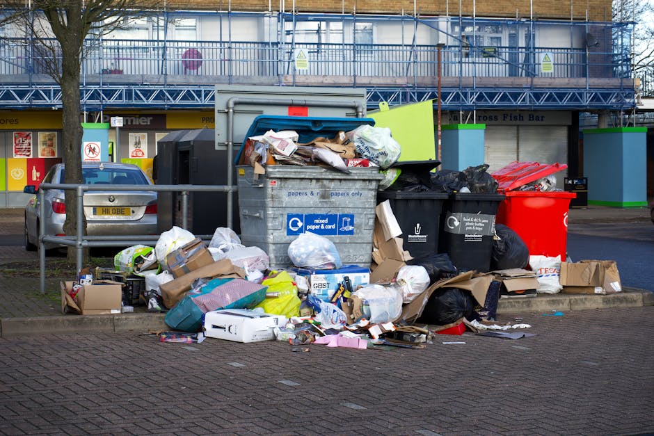 In a designated urban rubbish collection area, a large metal skip with a blue lid is overflowing with various types of waste, including crumpled brown paper, white plastic bags, cardboard boxes, and m