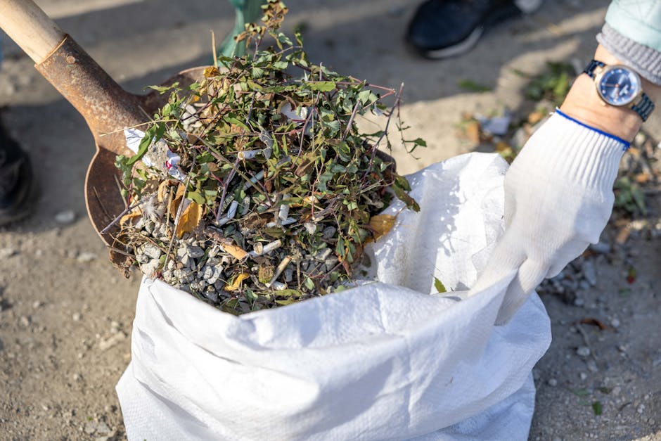 In a designated urban rubbish collection area, a large metal skip with a blue lid is overflowing with various types of waste, including crumpled brown paper, white plastic bags, cardboard boxes, and m