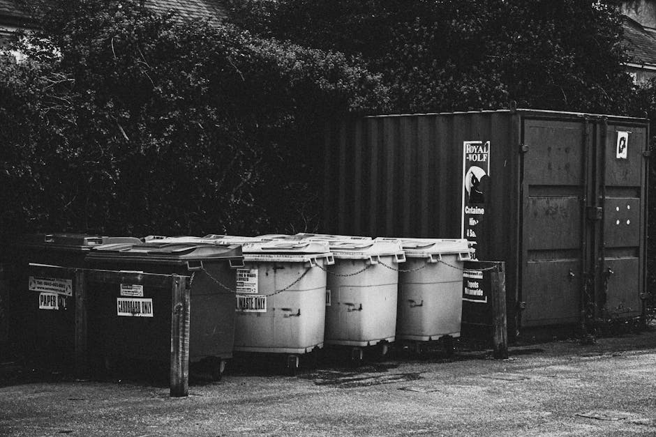A row of six large, white plastic wheelie bins with closed lids, positioned on a paved surface in front of a dark-colored shipping container. The bins are aligned side by side, with some labels indica